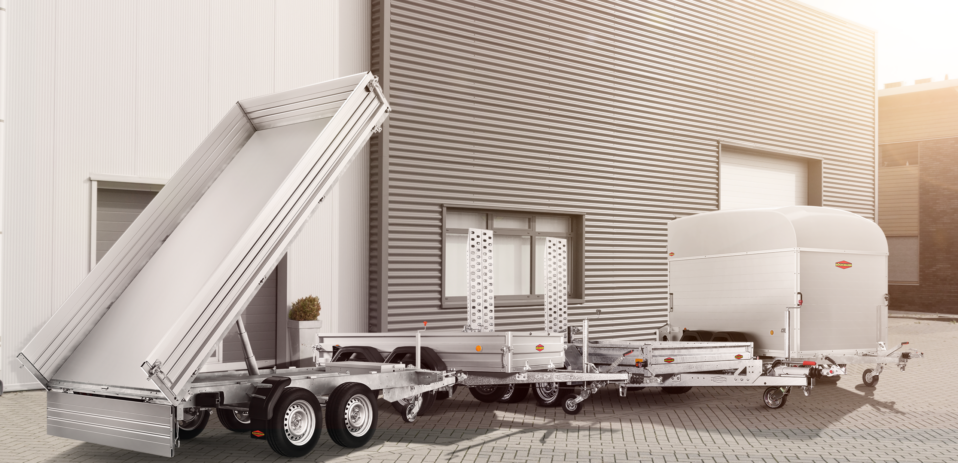 Vor einem modernen Industriegebäude mit Metallverkleidung und großen Toren steht eine Reihe von silbernen Nutzanhängern, darunter ein Kippanhänger, ein Tieflader und ein geschlossener Anhänger.