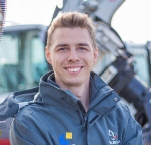 Ein junger Mann mit kurzen hellbraunen Haaren lächelt in die Kamera und trägt eine graue Jacke mit bunten Logos. Im unscharfen Hintergrund sind Baumaschinen zu sehen.