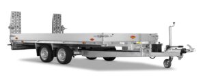Ein silberner Flachbett-Nutzfahrzeuganhänger mit zwei Achsen, einem vorderen Stützrad und zwei Metallrampen am Heck, der für den Transport von Fahrzeugen oder schweren Geräten ausgelegt ist.